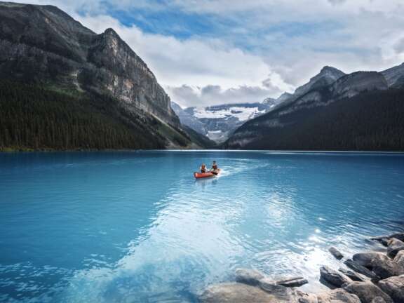 Two people canoeing in a red canoe on beautiful Lake Louise. The water is a vibrant blue and the mountains are glorious in the background.